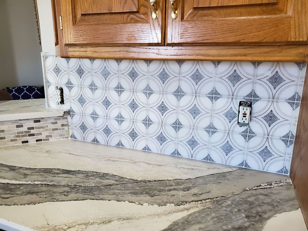 Kitchen backsplash of patterned white and gray tiles, with wood cabinets above and a speckled countertop below