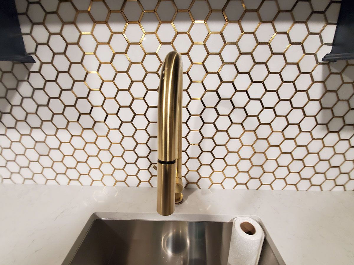 Gold faucet over a stainless steel sink, with honeycomb-tiled backsplash, and a roll of paper towels