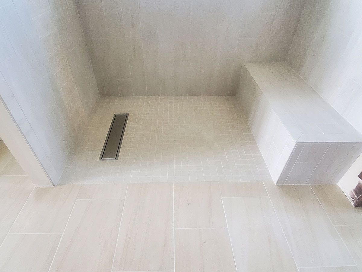 Beige tiled bathroom with bench, rectangular floor drain, and white walls