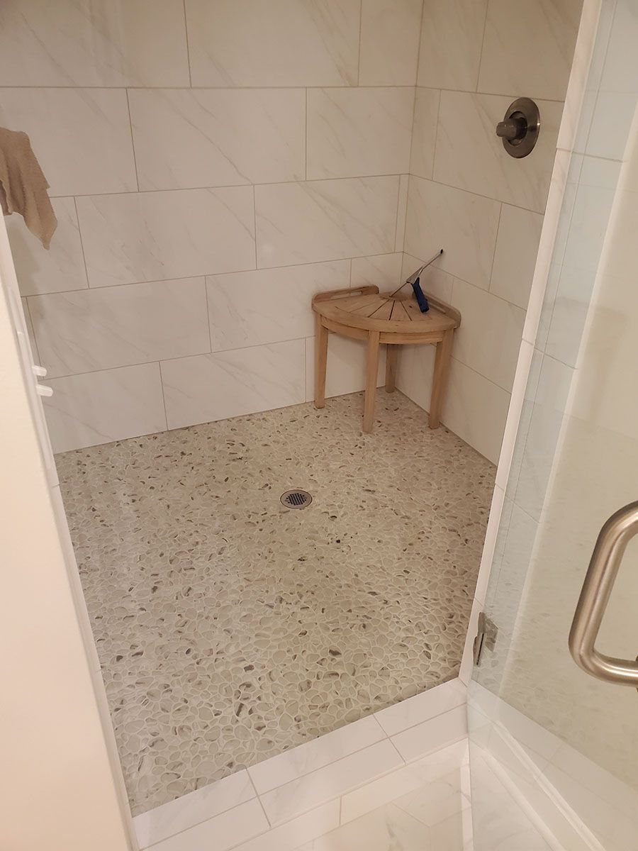 Shower with light-colored tile walls, speckled floor, and a small wooden stool