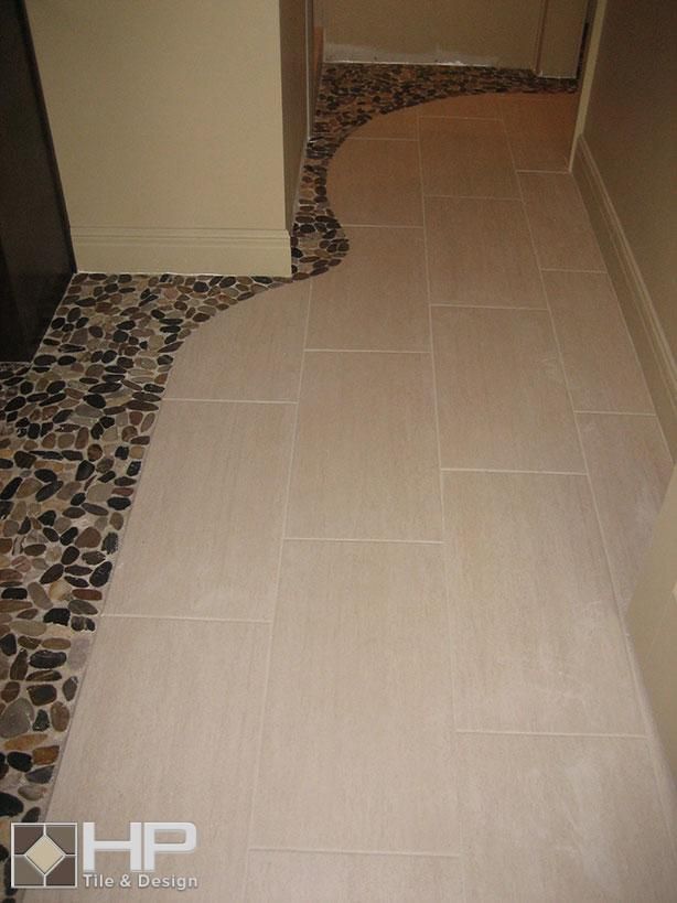 Floor with large, light-colored tiles meeting a pebble tile section with a curved edge; near a wall