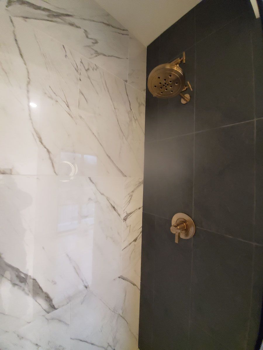 Shower stall with marble-patterned white wall and dark gray tiled wall; gold shower head and controls