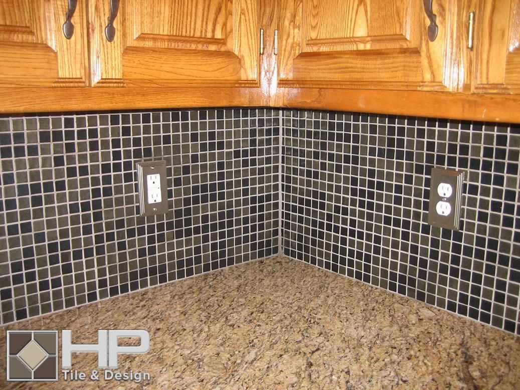 Kitchen backsplash with small black and beige tiles, granite countertop, and wooden cabinets.