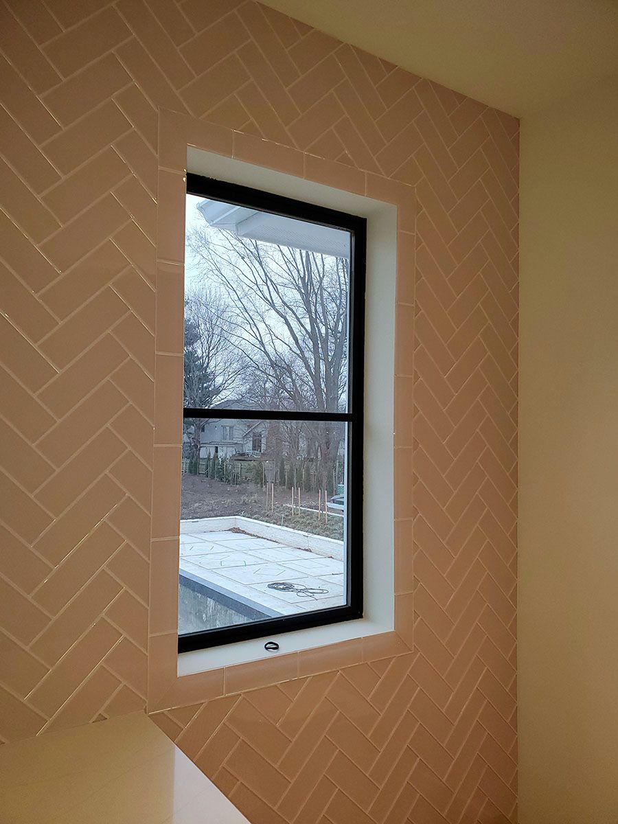 Tan herringbone tile wall with black-framed window overlooking a yard and pool