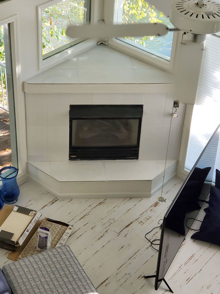 White tiled corner fireplace with black glass and white walls, in a room with a white ceiling fan and natural light