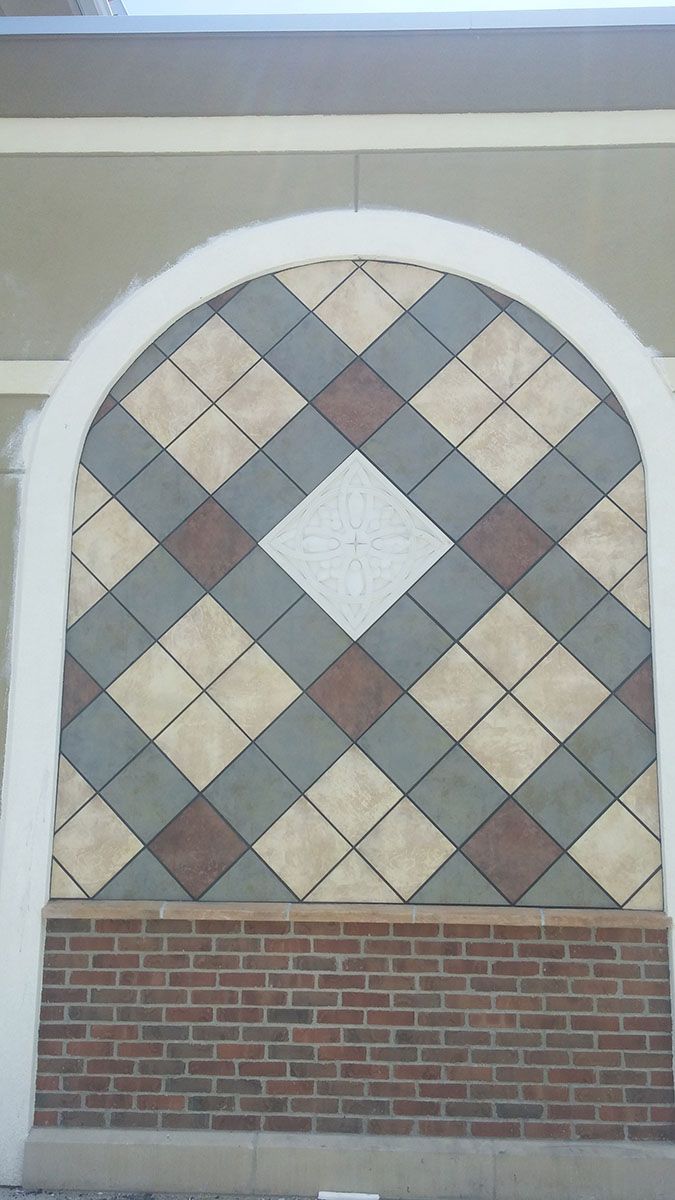 Decorative wall with diamond pattern using brown, gray, and beige tiles within an arched frame above a brick wall section