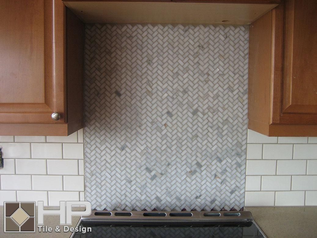 Kitchen backsplash with herringbone-patterned mosaic tile over stovetop, cream-colored subway tile below