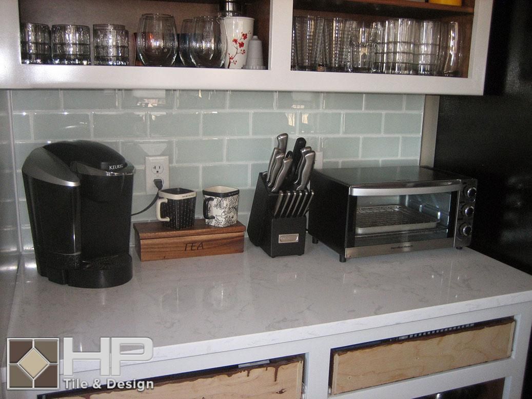 Kitchen counter with coffee maker, mugs, knife block, and toaster, against a light blue tile backsplash.