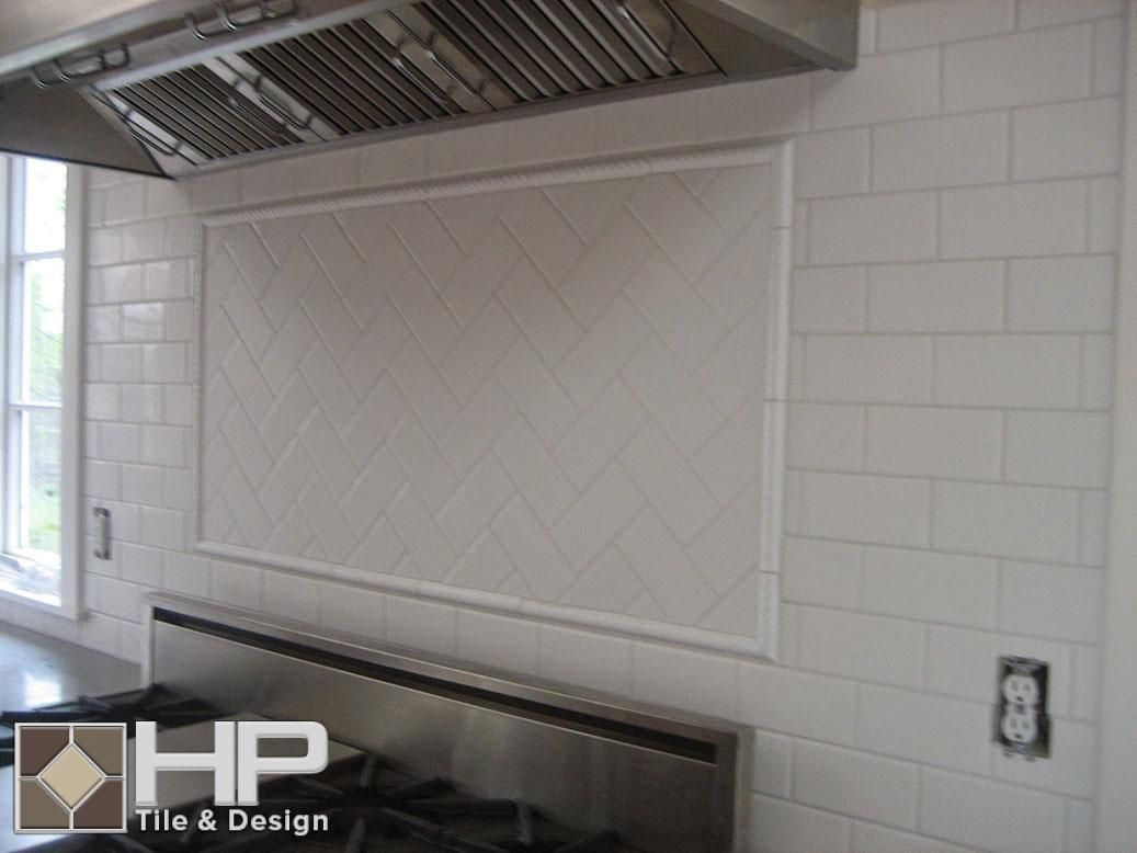 White tiled kitchen backsplash with a herringbone pattern, range hood, and stovetop.