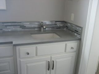 White cabinets with a gray countertop, white sink, and backsplash with a mixed tile pattern in a bathroom setting.