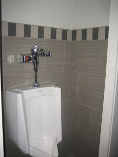 Urinal in a public restroom, white with chrome fixtures, gray tiled walls with a dark striped border
