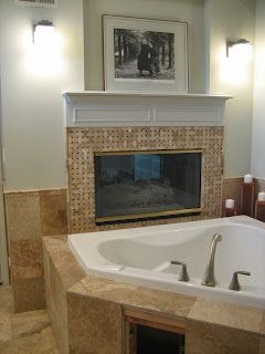 Bathroom with bathtub, fireplace, and artwork. Beige tile, white trim, two sconces.