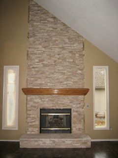 Stone fireplace with wooden mantel, flanked by tall, narrow windows, on beige wall.