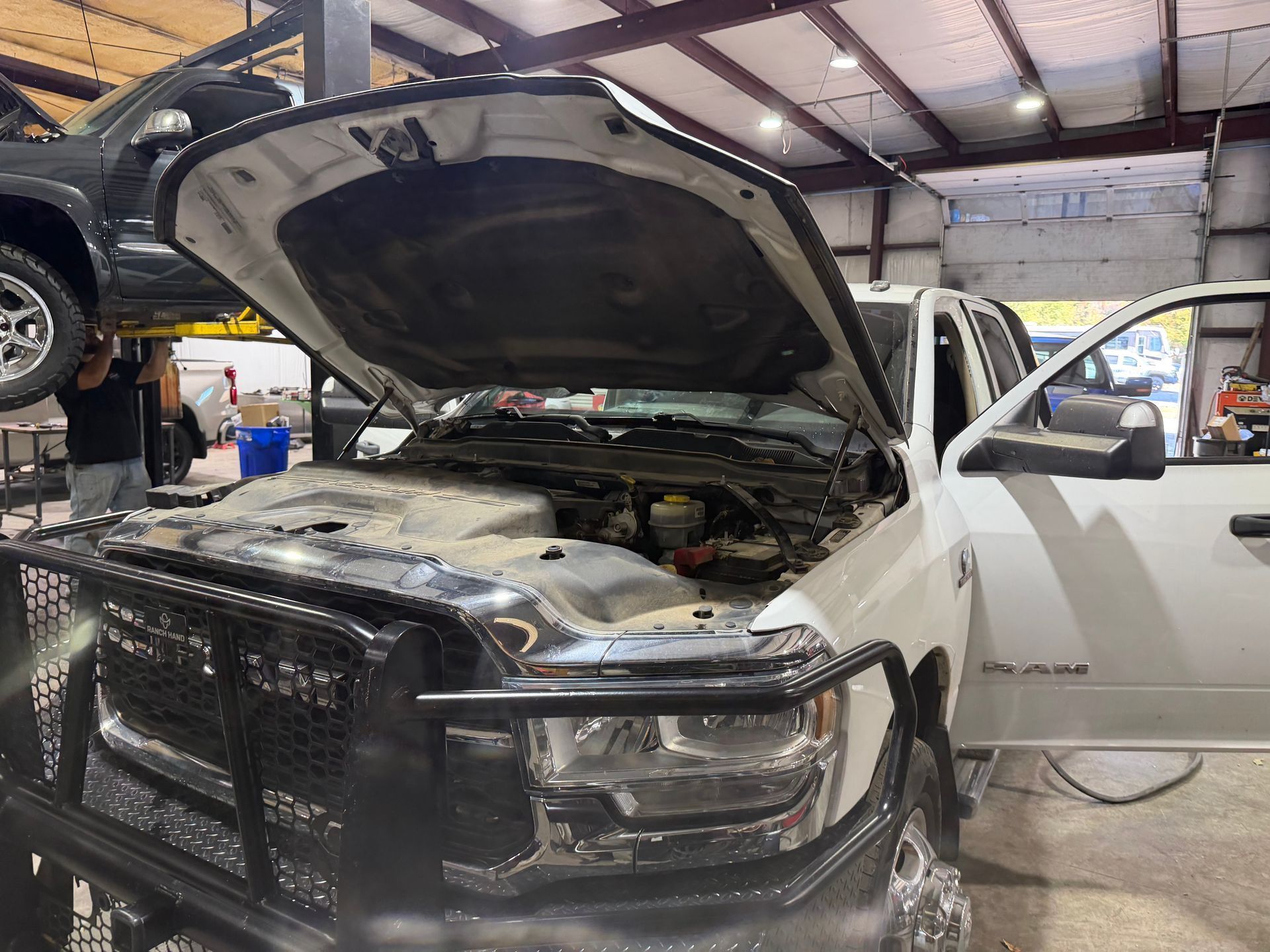 White pickup truck in a repair shop with hood open and front end damaged, mechanic working nearby