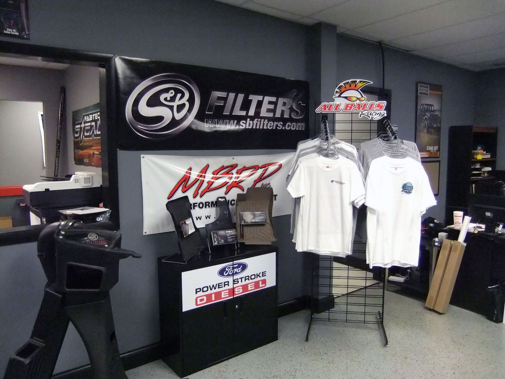 Trade show booth with car parts banners, white T-shirts on display, and a counter in a gray room