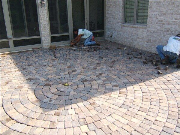 Two men are working on a brick patio with a circular pattern