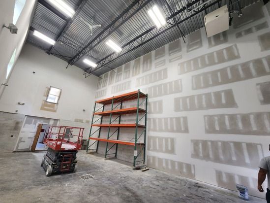 Warehouse interior with drywall, metal shelving, and a scissor lift under bright ceiling lights