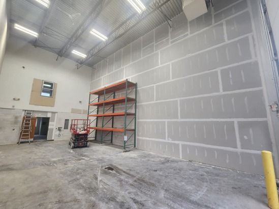 Empty industrial warehouse with high ceilings, drywall walls, metal shelving, and a pallet jack on the floor