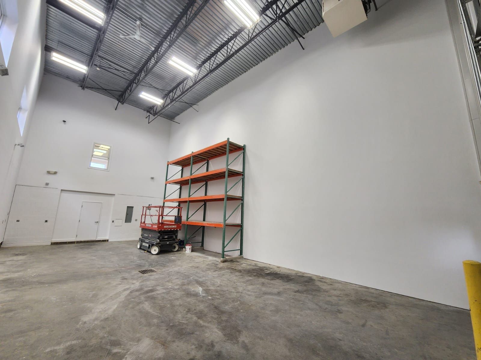 Empty warehouse interior with white walls, high ceilings, and metal shelving near a forklift stacker.