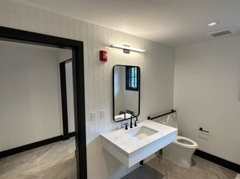 Accessible restroom with white sink, mirror, grab bars, and toilet in a modern hallway.