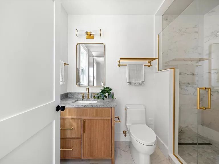 Bathroom with white walls, wood vanity, marble countertops, and gold accents. A glass shower enclosure and a toilet are also visible.