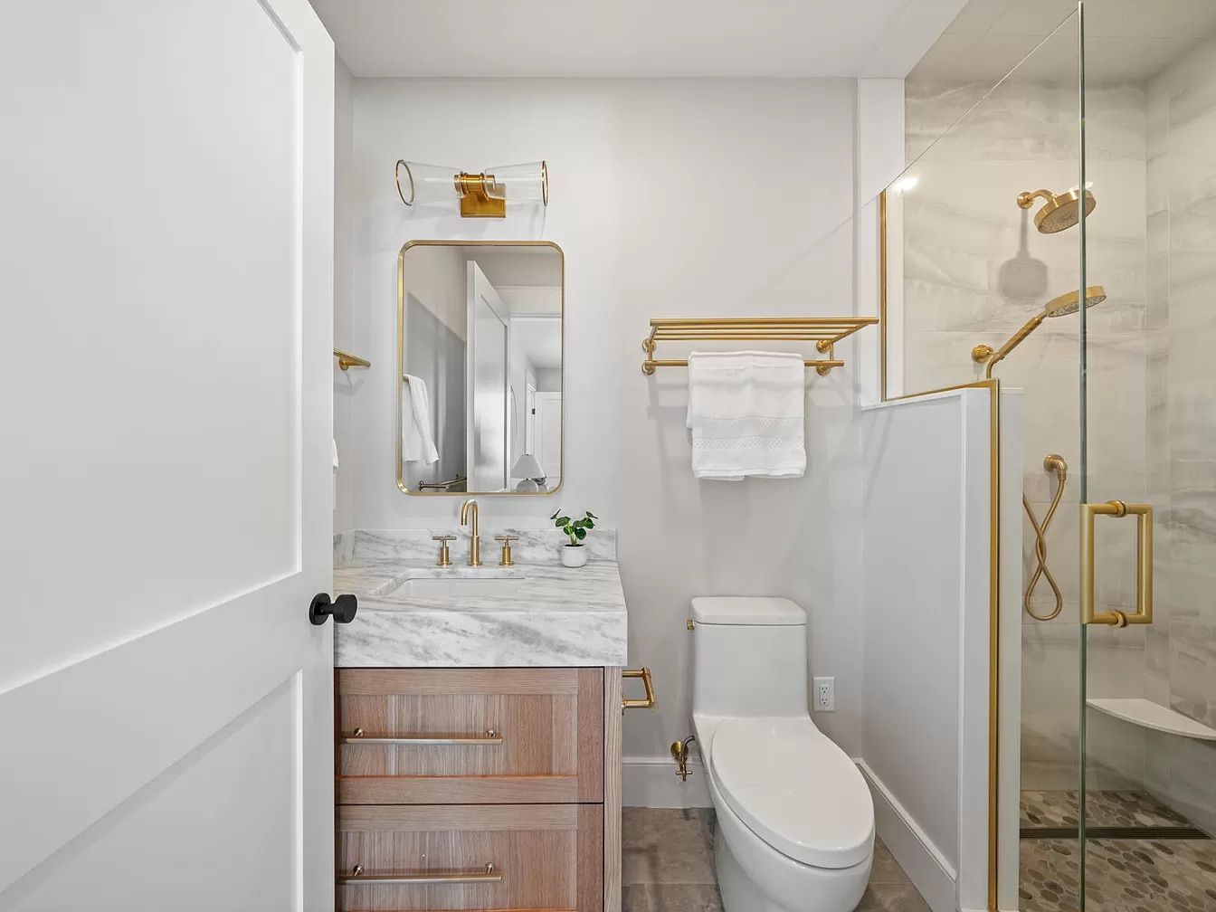A small bathroom with a vanity, toilet, and glass shower. Gold fixtures and a wood-tone vanity contrast with the light walls.