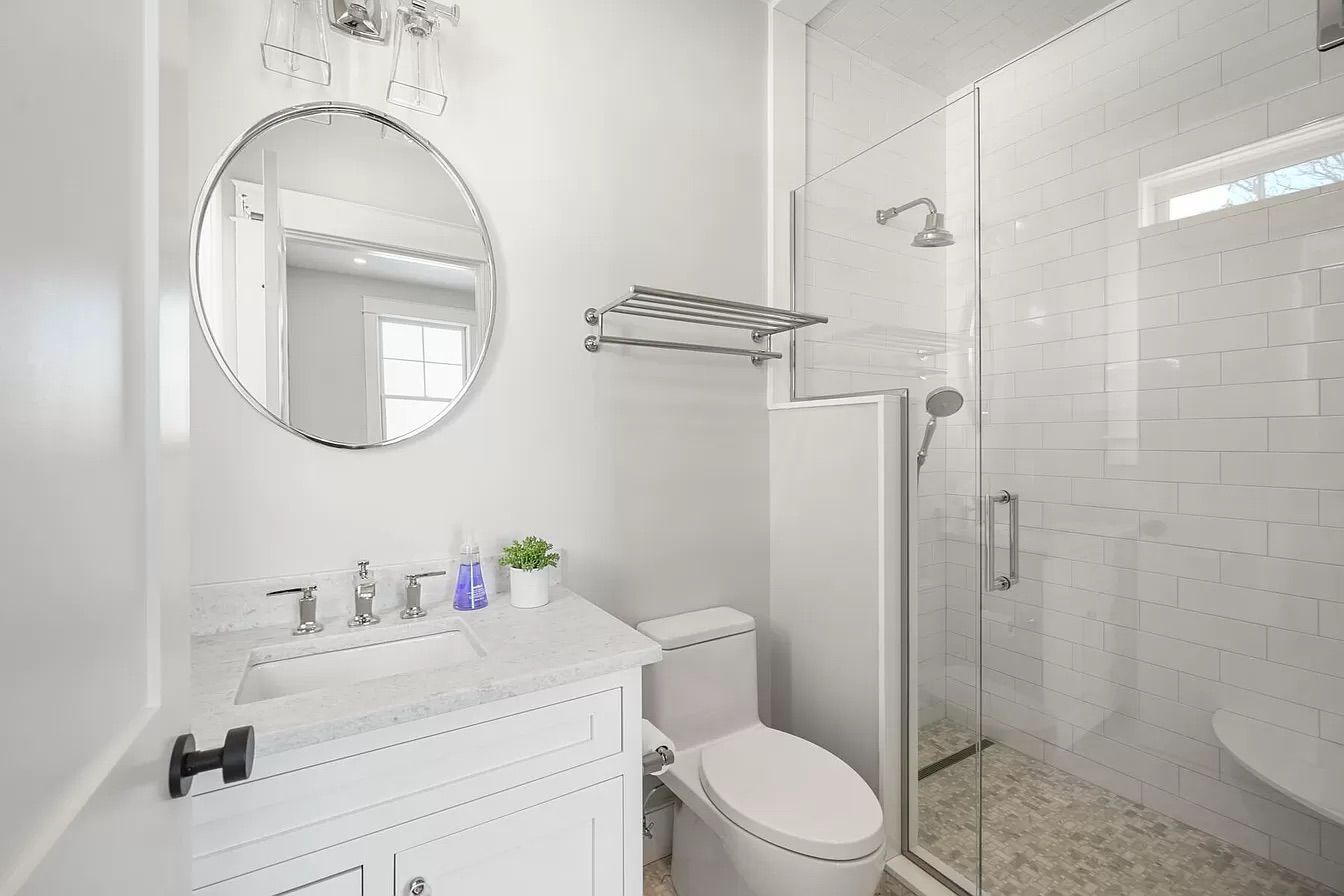 A bright white bathroom with a glass-doored shower, toilet, sink, and round mirror.