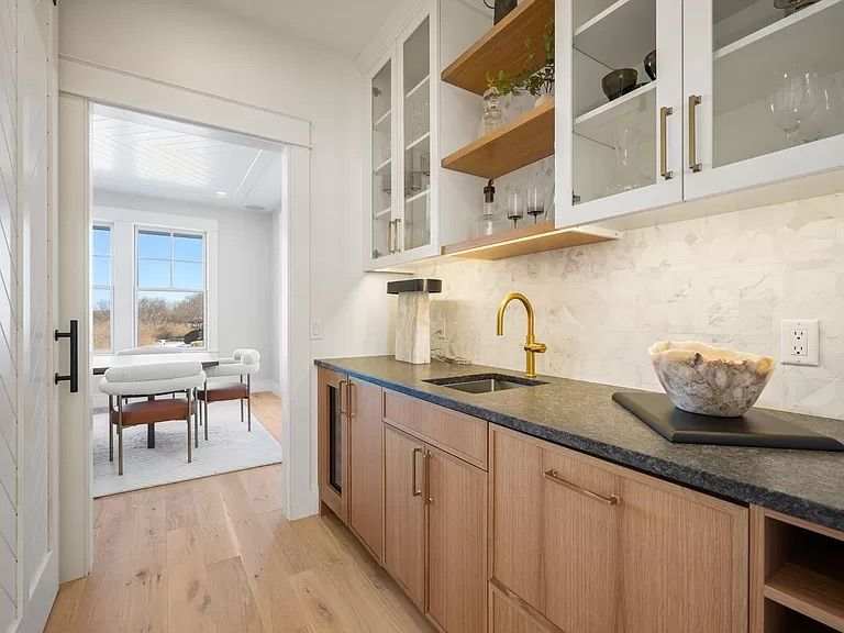 A modern butler's pantry with light wood cabinets, a dark countertop, and a brass faucet. A doorway leads to a bright dining area.