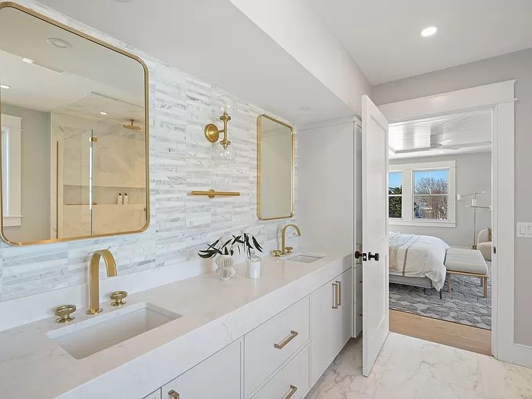 A modern bathroom with white countertops, gold fixtures, and marble accents. A doorway leads to a bedroom with a bed and rug.