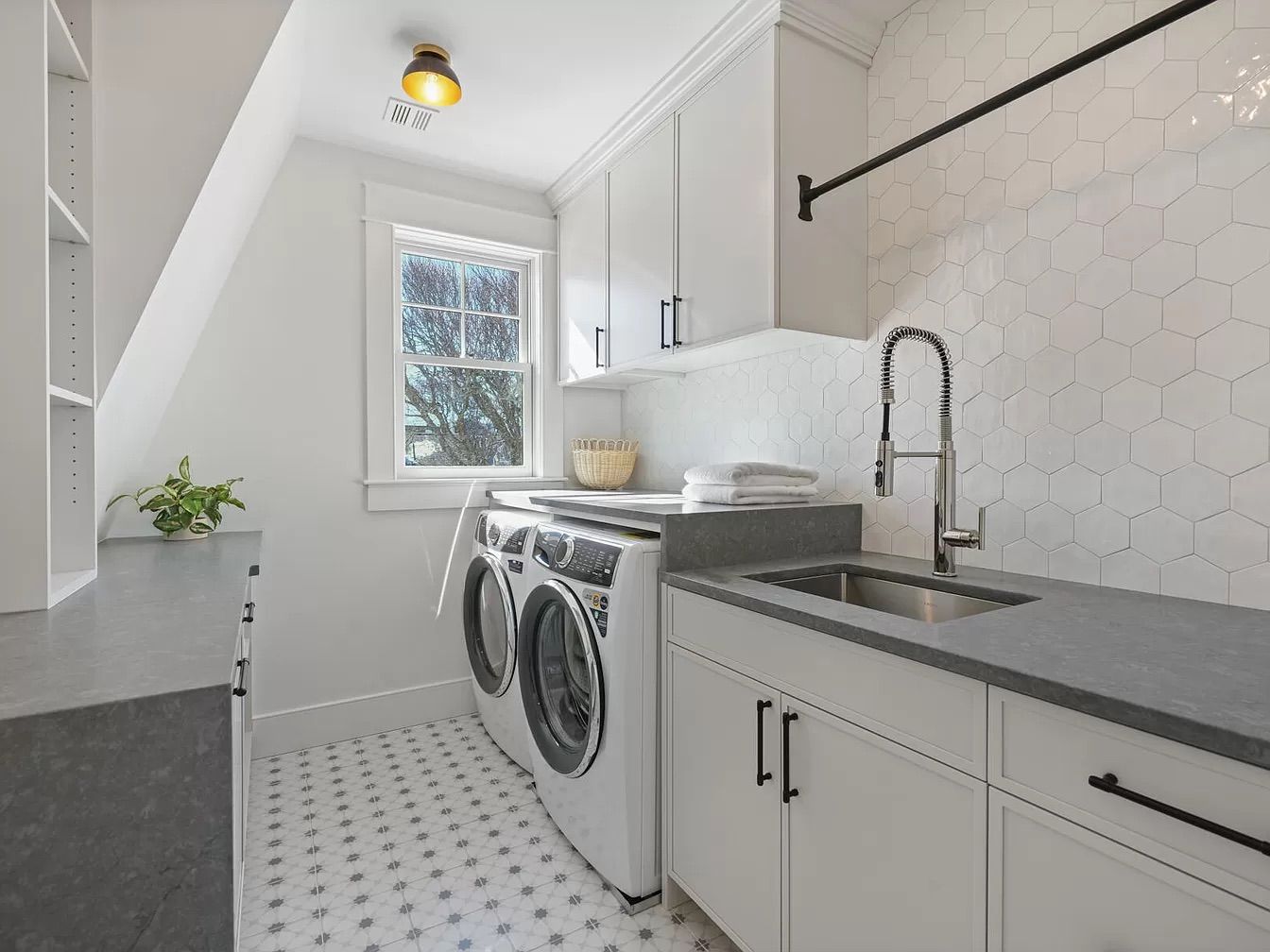 A well-lit, modern laundry room with white cabinetry, a washing machine and dryer, a utility sink, and honeycomb-patterned tile backsplash.