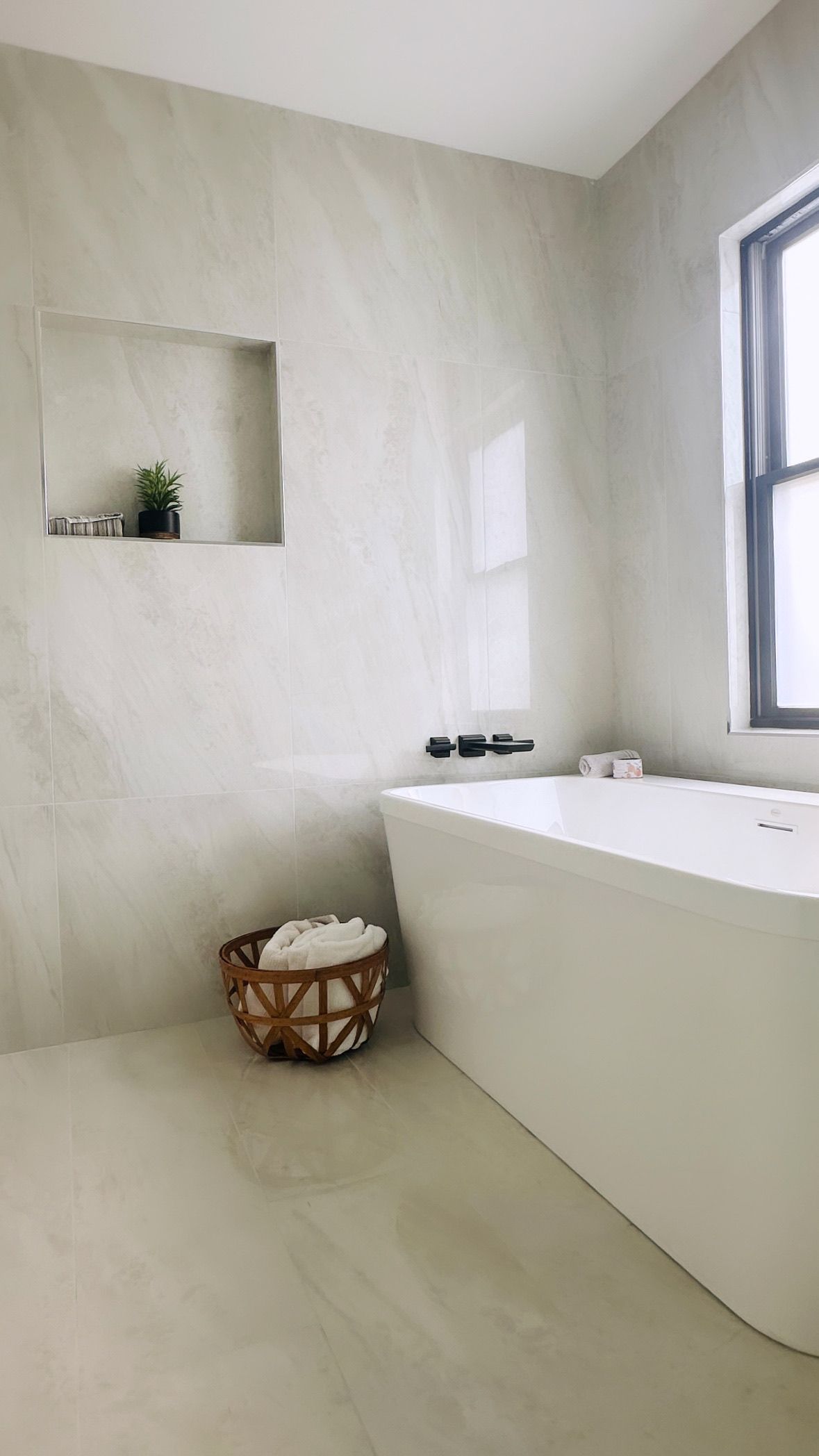 Modern bathroom with a white bathtub, neutral-toned walls, and a wicker basket holding towels. A small plant sits in a recessed shelf.