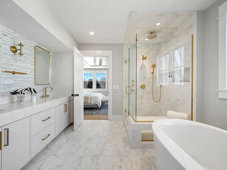 Modern bathroom with white marble tile, gold fixtures, a glass shower, and a view into a bedroom through an open door.