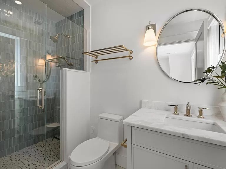 Modern bathroom with a glass shower, white vanity, and round mirror. The walls are white, and the shower has dark blue-green tiles.