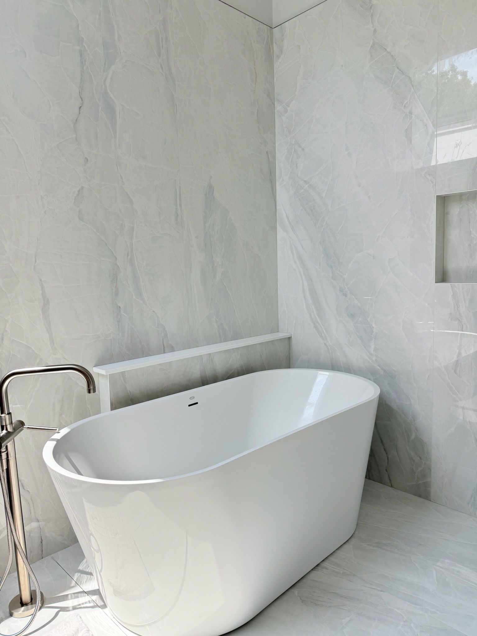A modern bathroom with a white freestanding bathtub. Gray marble-like tiles cover the walls and floor.