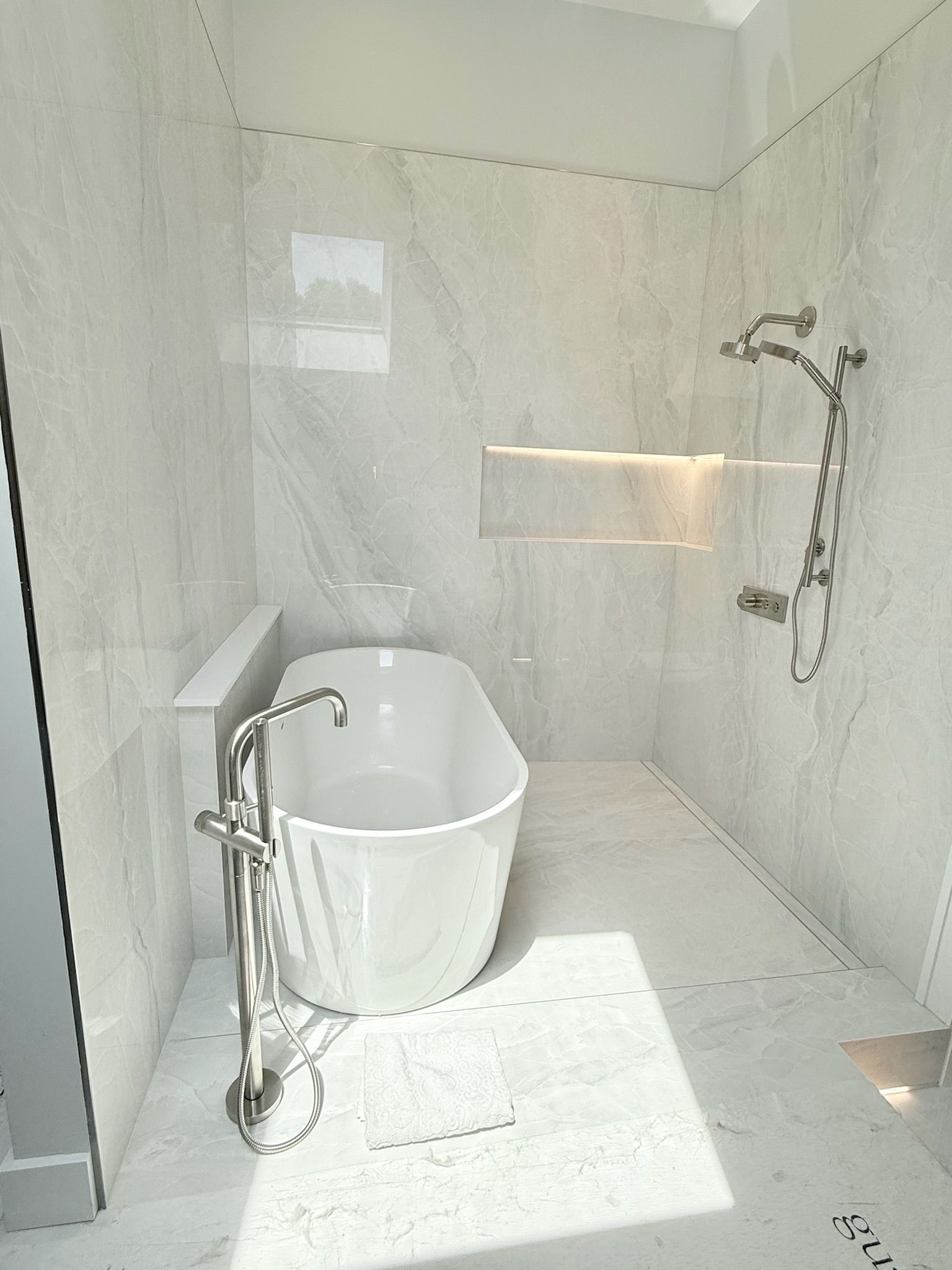 Bathroom with a white soaking tub, shower fixtures, and light-colored stone tile. The room is minimalist and bright.