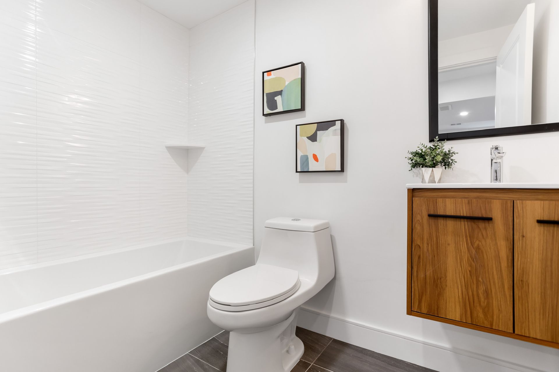 A modern bathroom with white textured tile, a toilet, a wood-grain vanity, and abstract art.