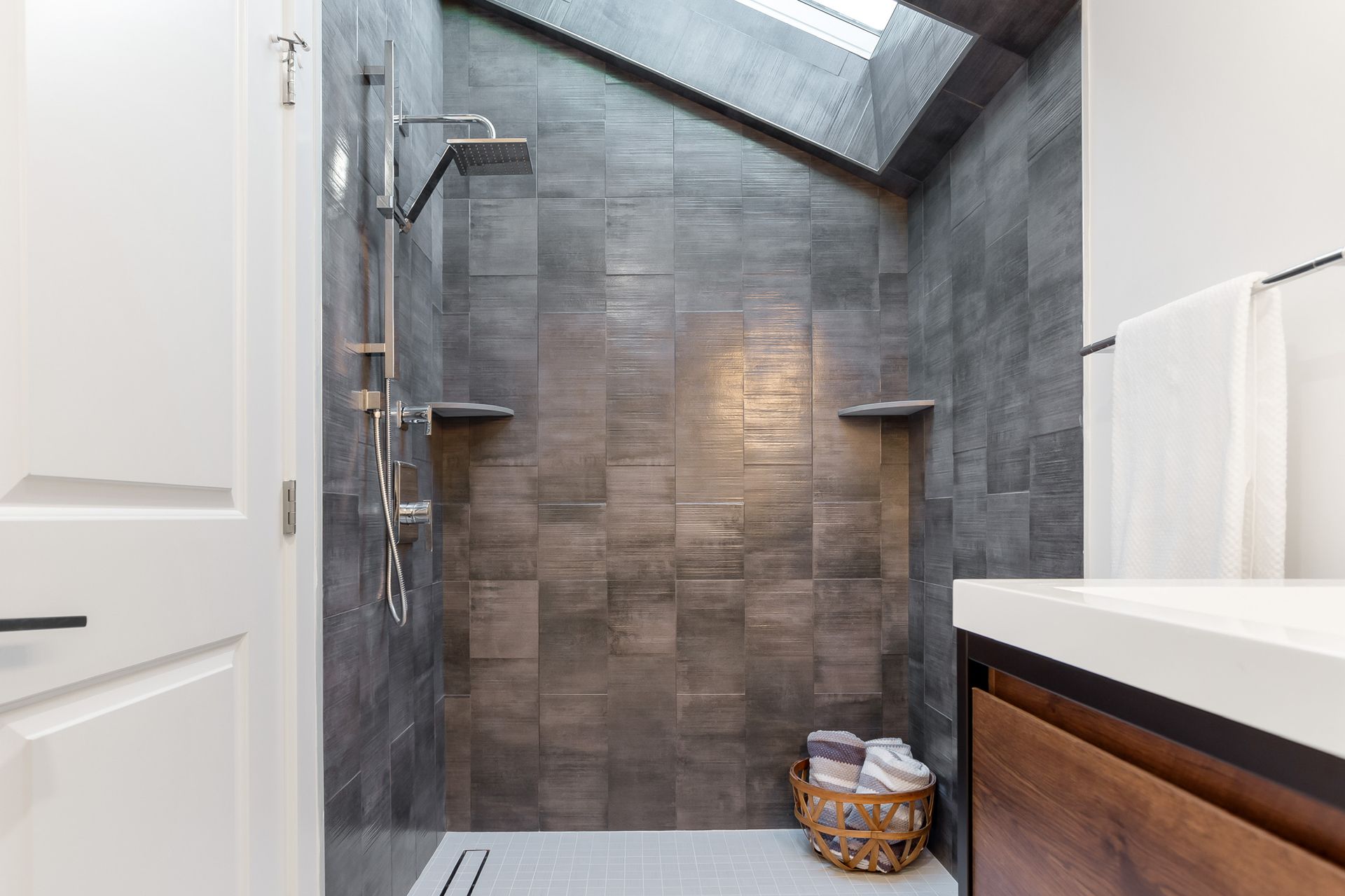 A modern bathroom with a dark tile shower featuring a skylight. A basket of towels sits near the vanity.