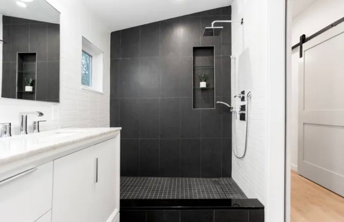 Modern bathroom with a black-tiled shower, white vanity, and a sliding barn door. A window lets in light.