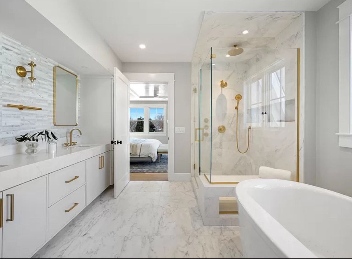 A luxurious bathroom with white marble tiles, a gold shower, and a bathtub. The open door reveals a glimpse of a bedroom.