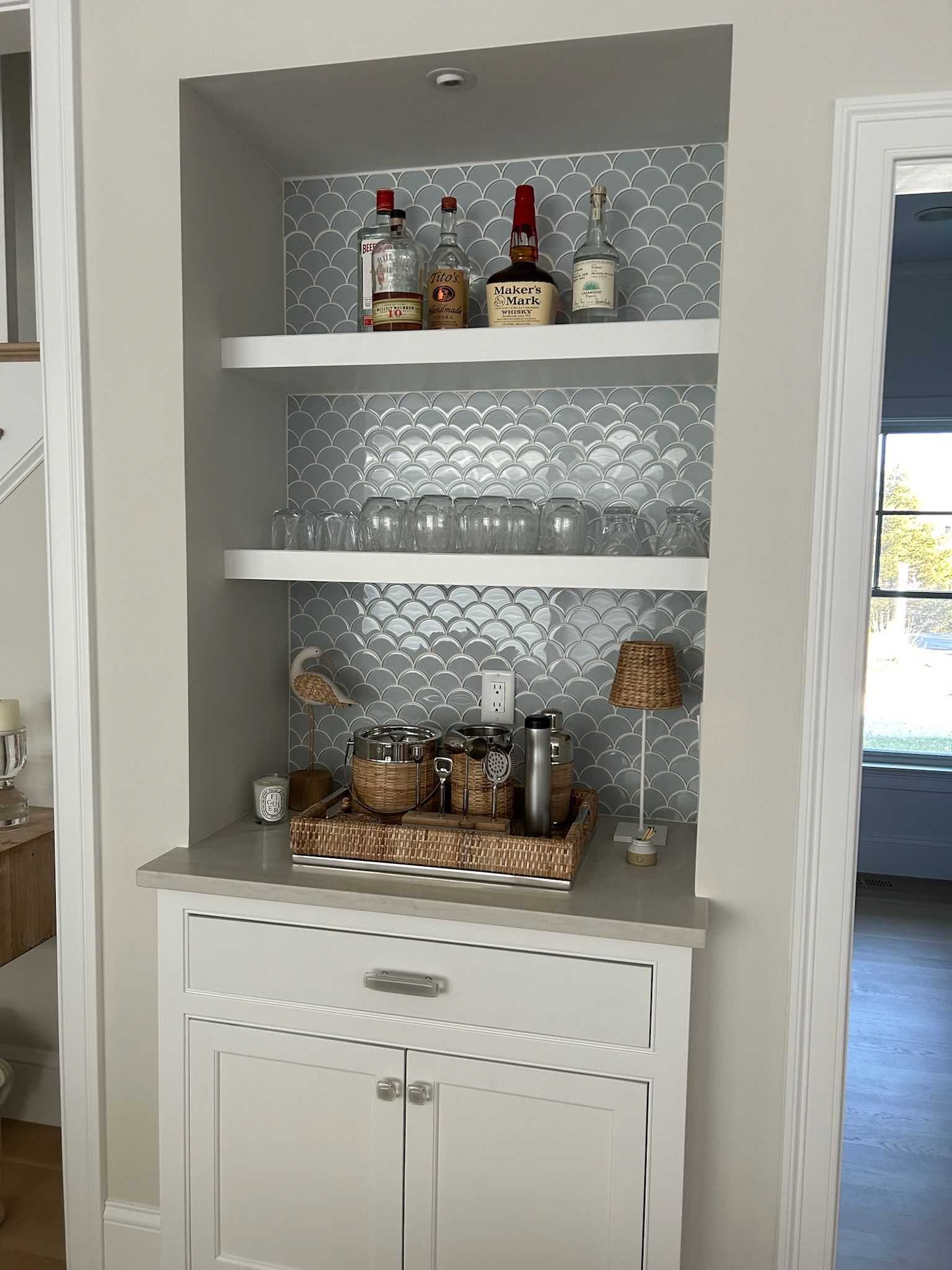 Built-in bar with white cabinetry, shelves, and decorative backsplash, stocked with liquor, glassware, and a woven tray.