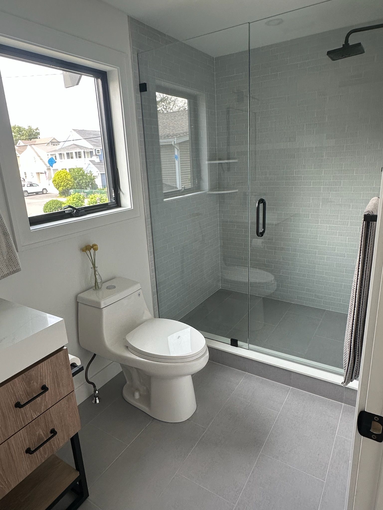 A modern bathroom with a toilet, vanity, and glass-enclosed shower with gray tile. A window provides a view outside.