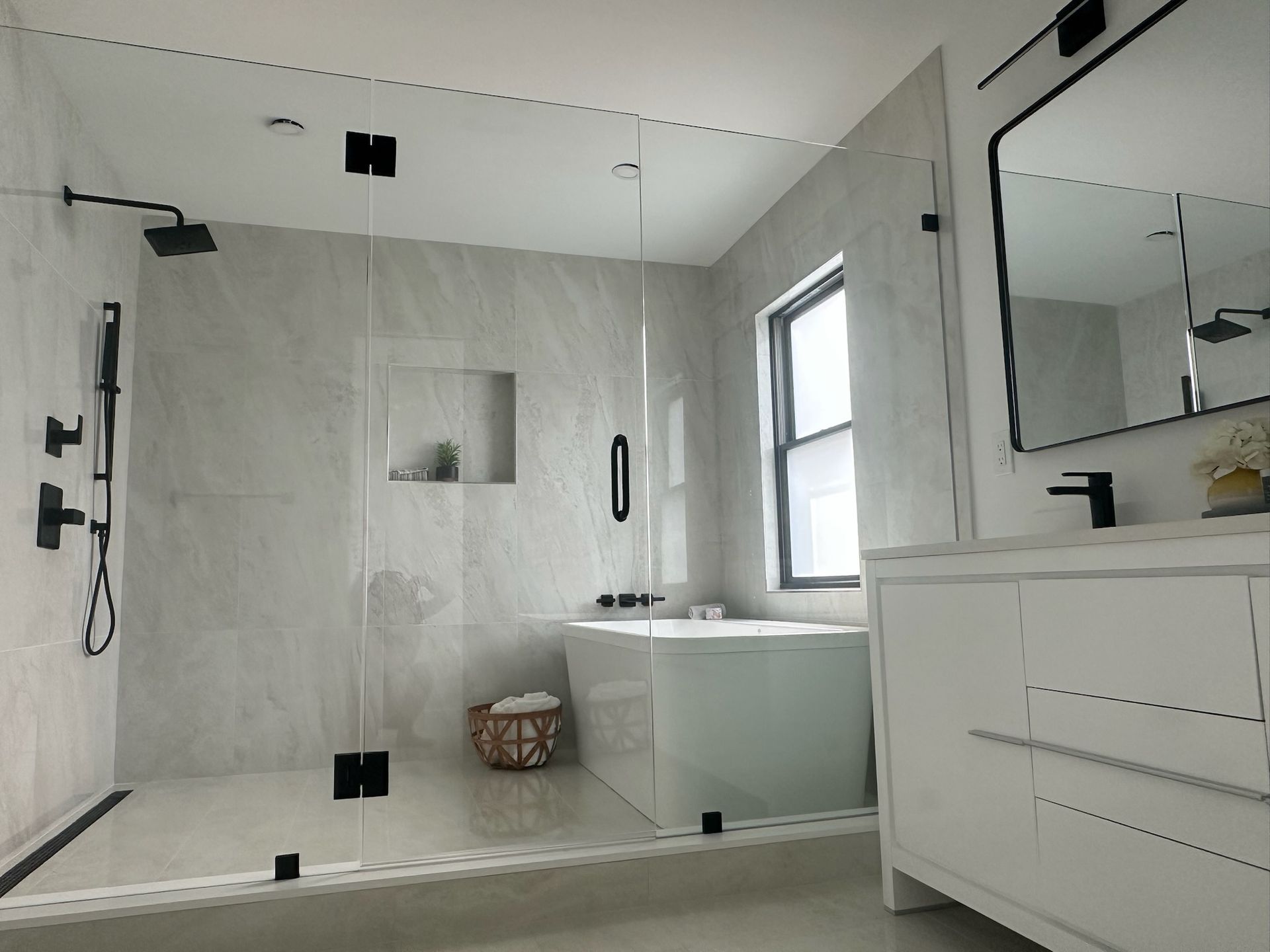 Modern bathroom with a glass shower, a soaking tub, a white vanity, and light gray tile. Black fixtures are visible throughout.