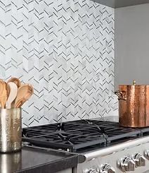 A kitchen backsplash with a white and gray herringbone pattern, with a copper pot on the stove. Wooden spoons sit in a canister.