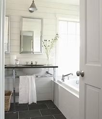 A modern, white bathroom with a glass-legged vanity, a large mirror, a bathtub, and a basket. Dark tile flooring contrasts with the light walls.