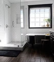 A modern bathroom with a black bathtub, shower, window, and dark wooden floors. The walls are tiled white.