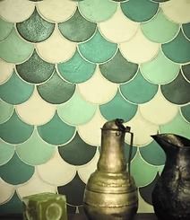 A wall with teal, green, and white scale-like tiles. In front sit a bronze pitcher, a black pitcher, and a green candle.