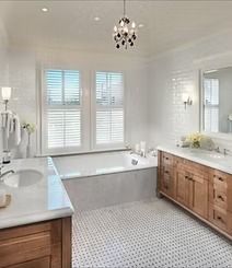 Spacious, bright bathroom with white subway tile, wood cabinets, a tub, and a chandelier. Windows with white shutters let in natural light.