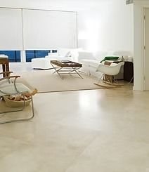 Bright living room with a white sofa, rug, chair, and a light-colored polished floor. Large windows with white shades.