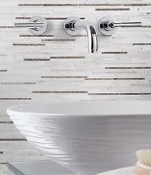 Wall-mounted chrome faucet and handles above a white, textured sink, against a white and gray mosaic tile backdrop.