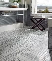 Bathroom with gray brick tile walls, a wooden stool, and a floor with gray and white linear tiles. Sunlight streams through the window.